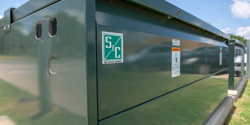 Closeup of one corner of S&C’s Vista Underground Distribution Switchgear. The green S&C logo is in the foreground. In the background is a blue sky with puffy white clouds.