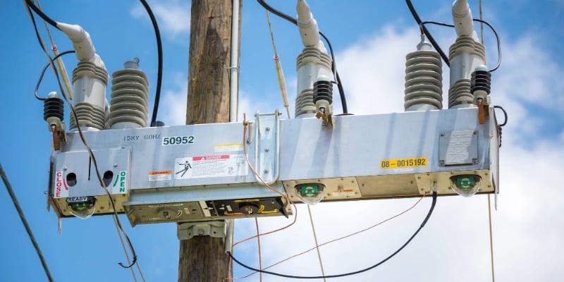 An S&C IntelliRupter® PulseCloser® Fault Interrupter rests on a pole in the foreground with electrical wires connected to it. In the background is a blue sky with wispy white clouds.